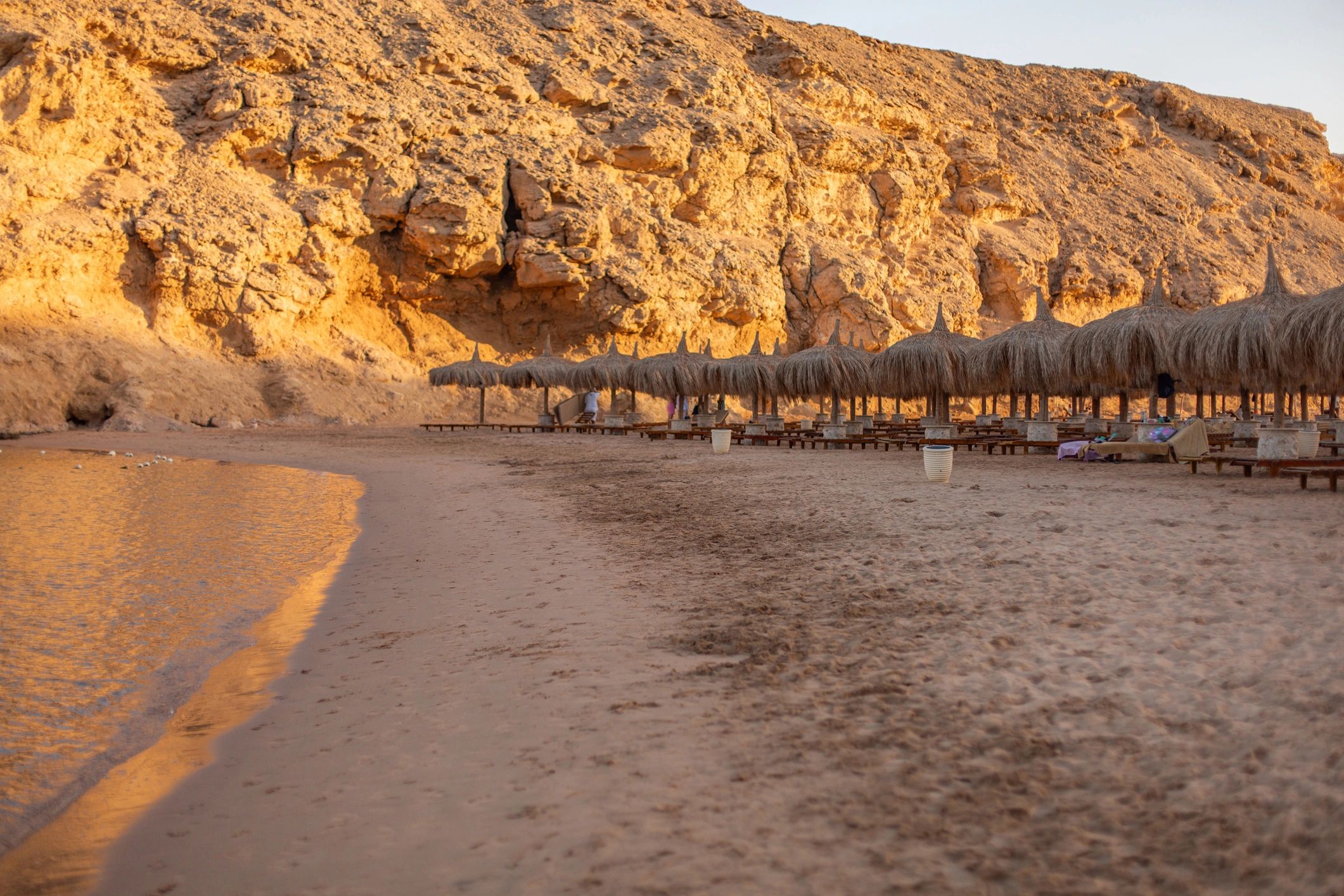 Sandy beach with straw umbrellas under desert mountains during sunset